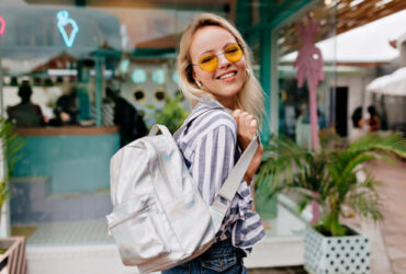 Mulher com look despojado de calça jeans, mochila, camisa listrada e óculos de sol