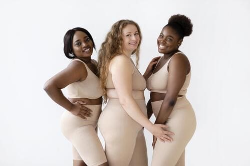 3 mulheres sorridentes posando com suas respectivas roupas intimas plus size no tom nude