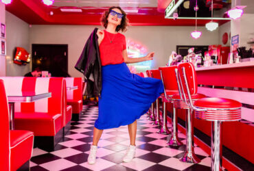 Mulher usando um look dos anos 60 com blusa vermelha vestido azul e óculos escuro